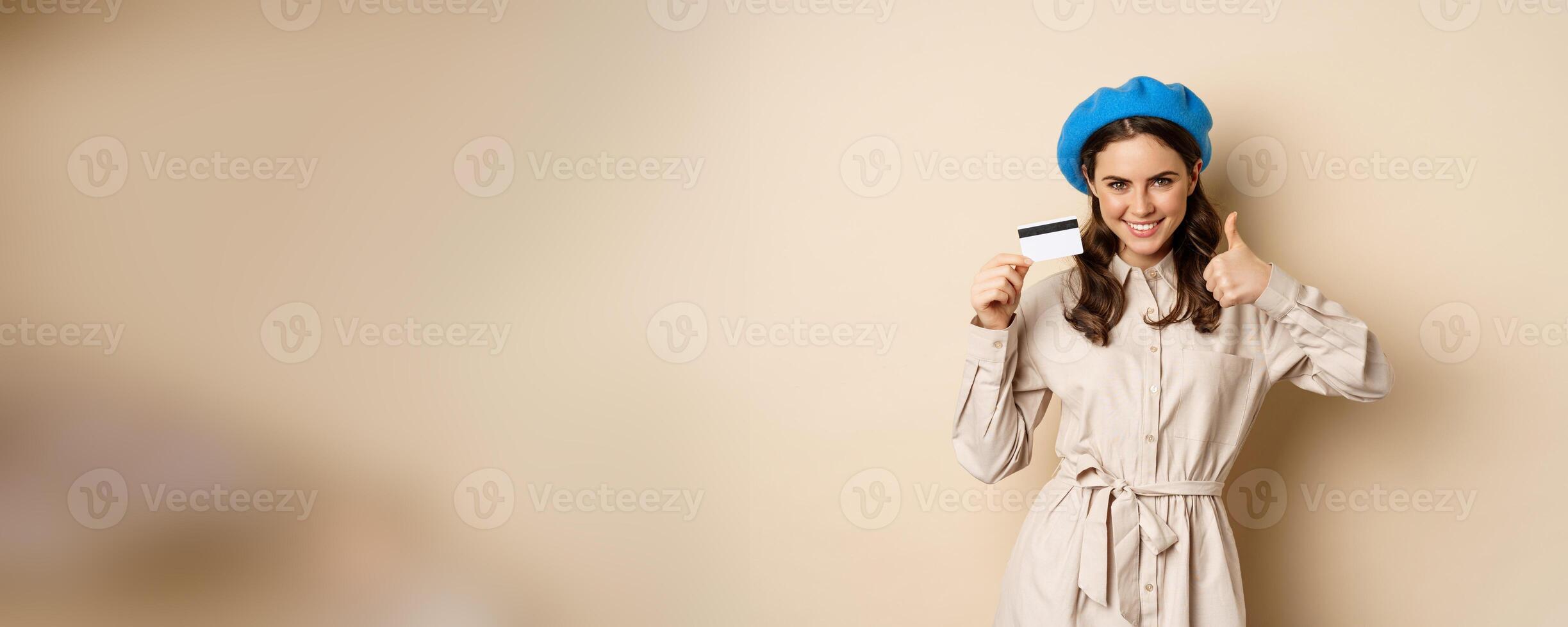 Concept of shopping and sale. Happy young woman in stylish clothes, showing credit card store discount and thumbs up, recommending, standing over beige background photo