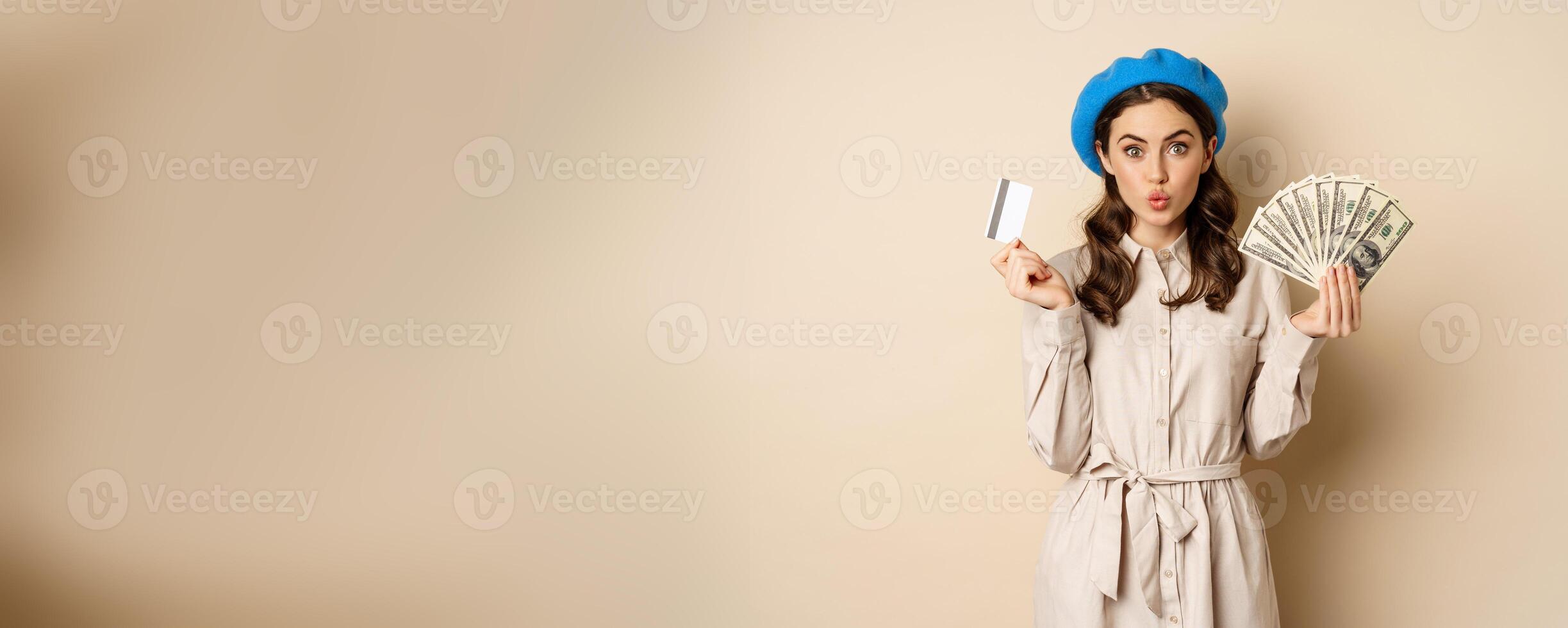 Microcredit and money concept. Young stylish woman showing credit card and dollars cash, smiling happy and satisfied, standing over beige background photo