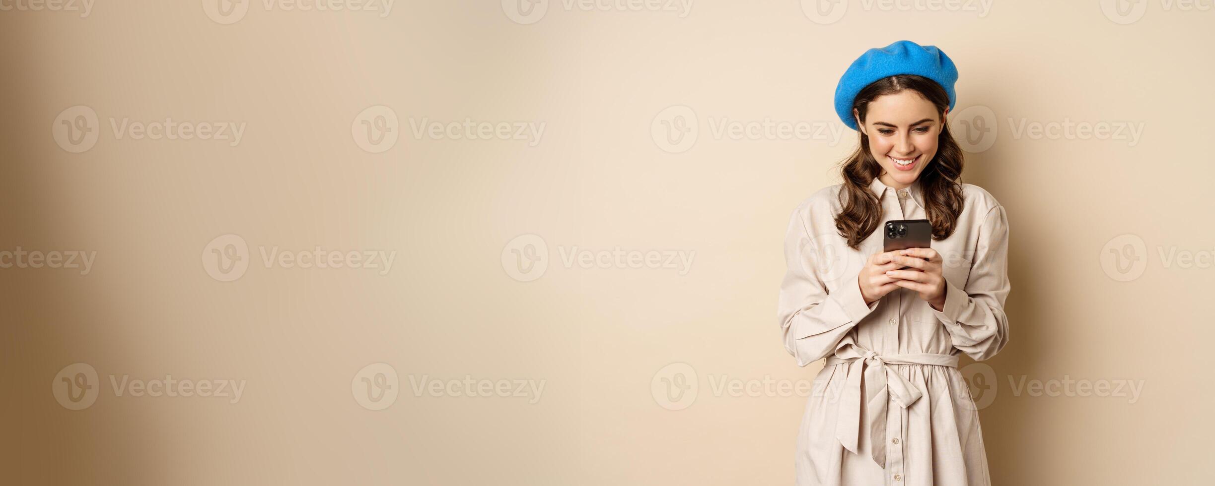 Modern girl using smartphone, smiling and posing happy with mobile phone, standing over beige background photo