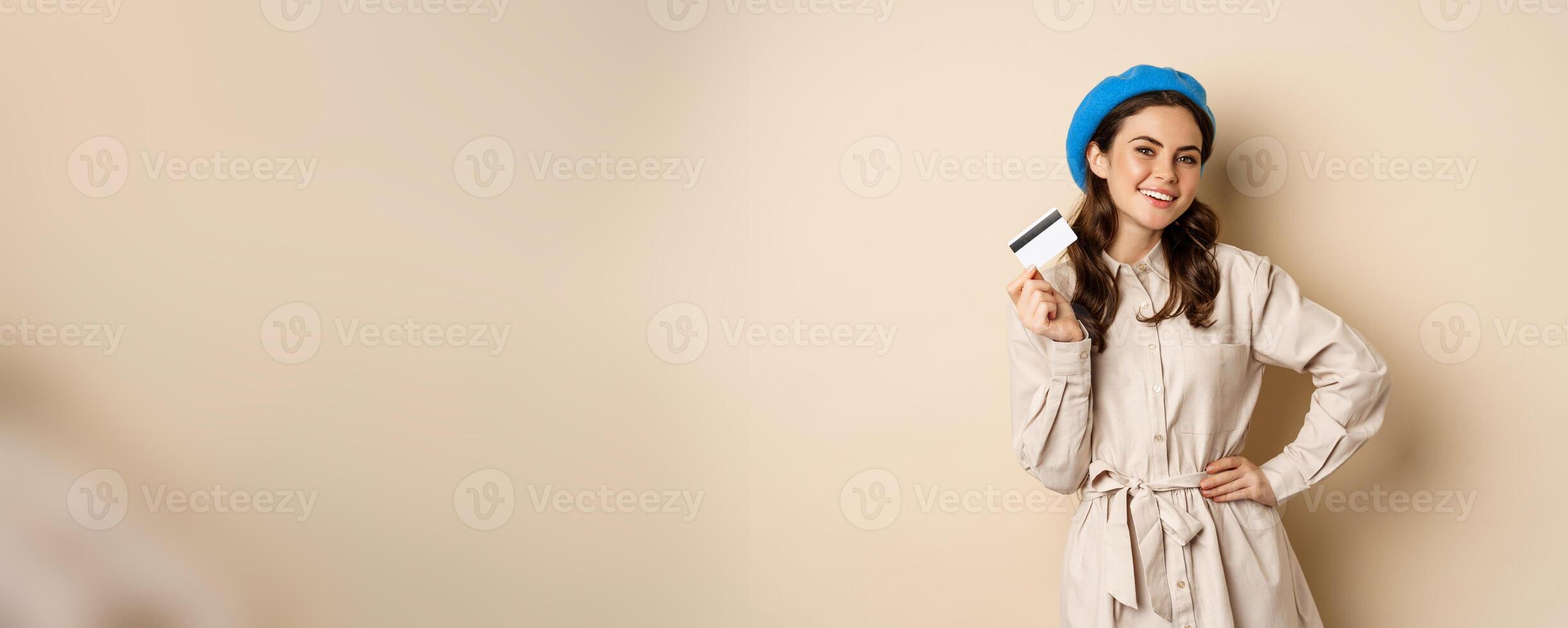 Shopping and discounts concept. Young stylish modern girl, posing with credit card, looking happy and smiling, standing over beige background photo