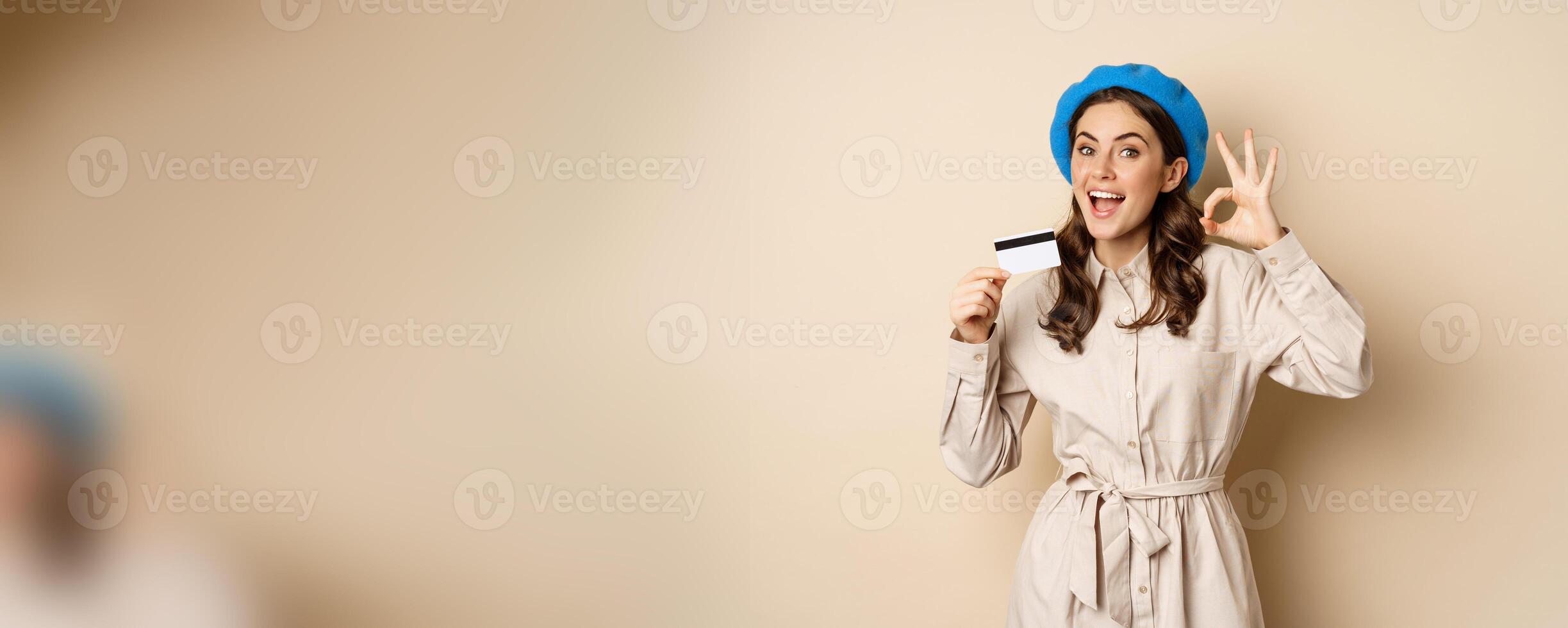 Concept of shopping and sale. Happy young woman in stylish clothes, showing credit card store discount and okay, satisfied, like and approve smth, standing over beige background photo