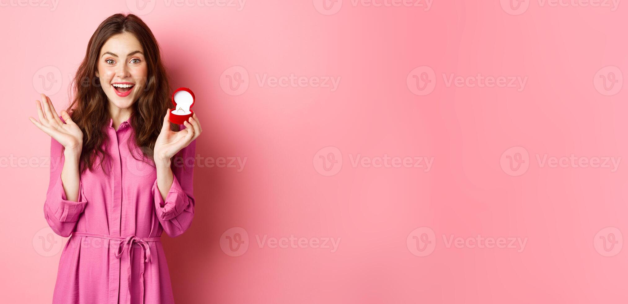 Excited young woman showing engagement ring, getting married, smiling happy, talking about wedding day, receive marriage proposal, standing over pink background photo