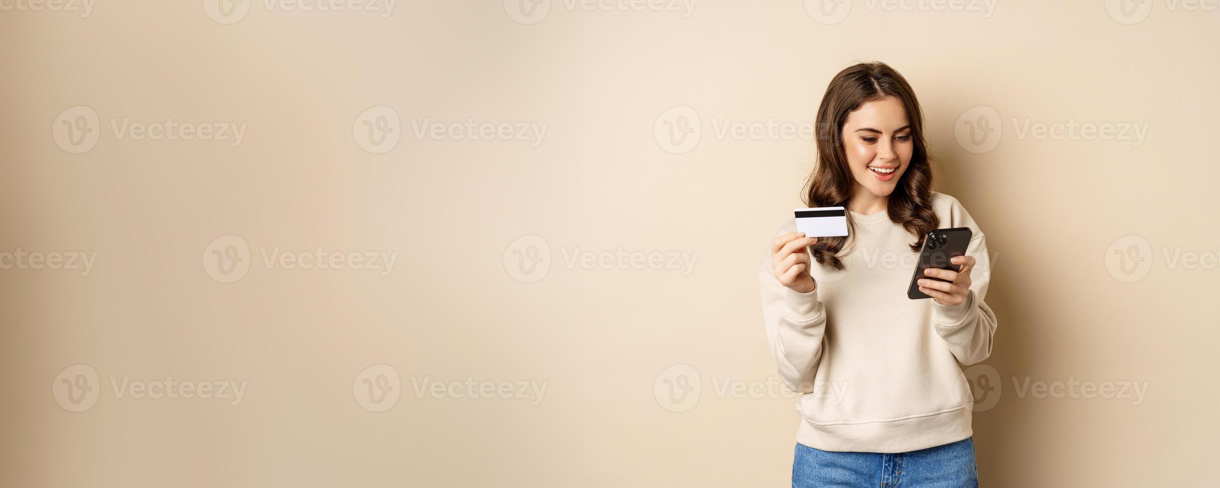 Smiling girl using mobile app, smartphone shopping and credit card, standing over beige background, order smth photo