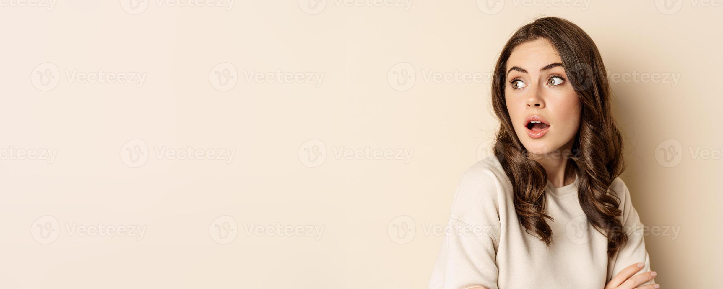 Close up portrait of brunette feminine woman turn back, look behind with surprised face expression, standing against beige background photo