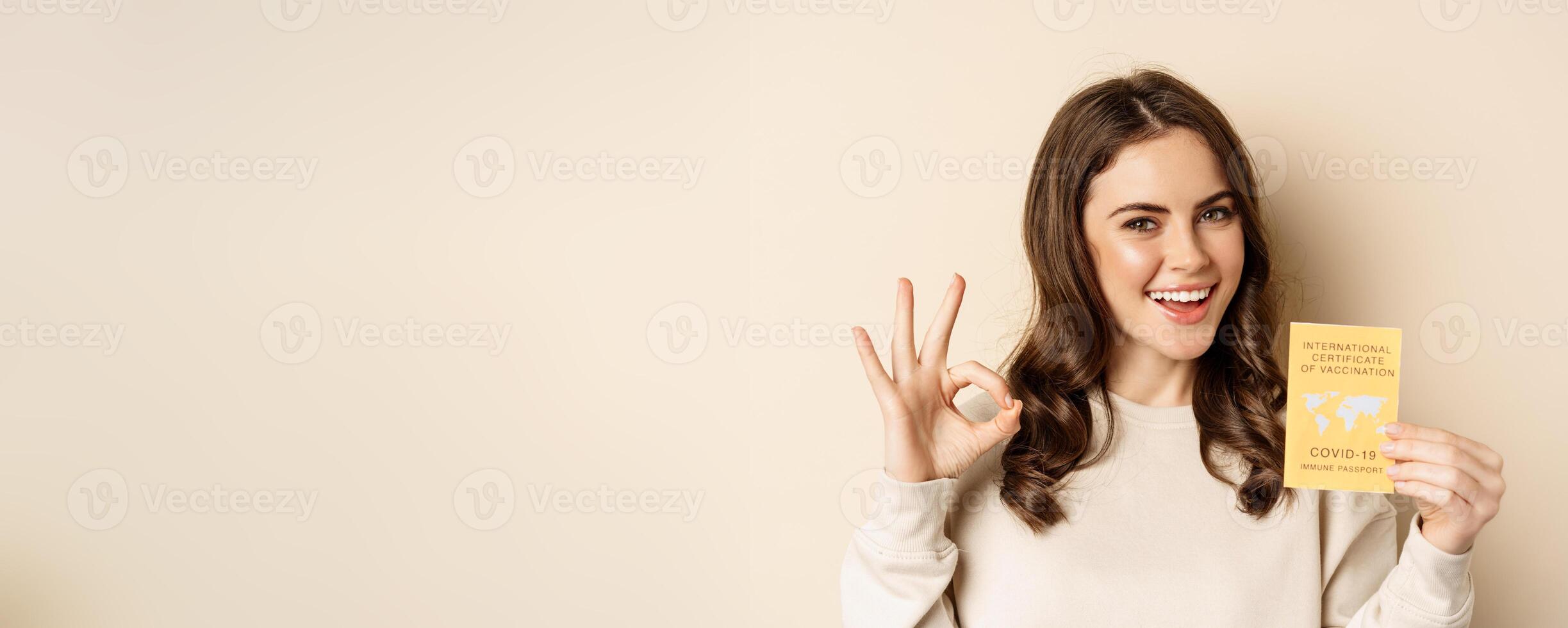 Travelling and pandemic concept. Portrait of woman showing international covid vaccination certificate, okay sign, easy travel pass, beige background photo