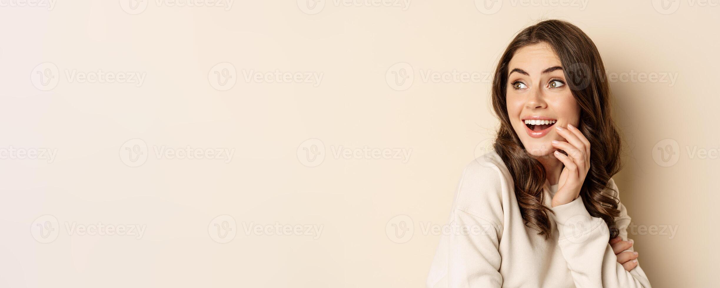 Close up portrait of brunette feminine woman turn back, look behind with surprised face expression, standing against beige background photo