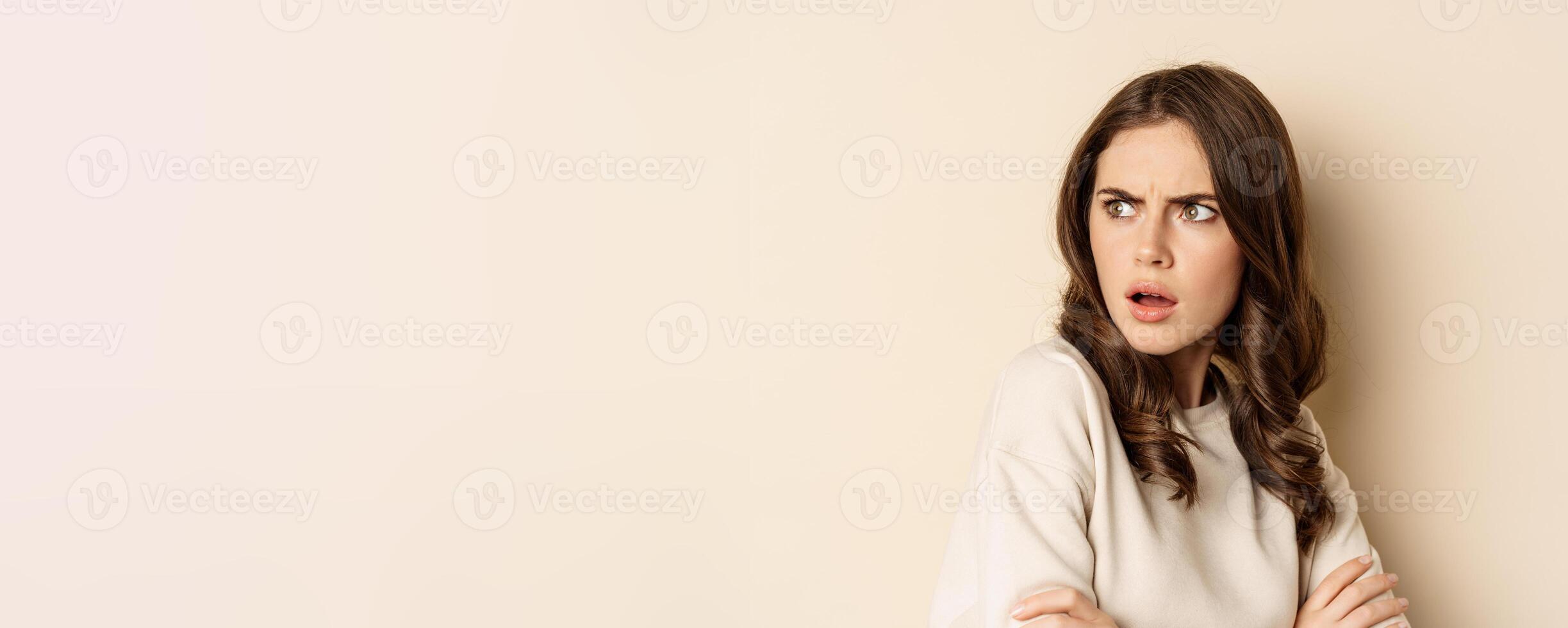Close up of confused young woman, looking behind with skeptical shocked face expression, standing against beige background photo