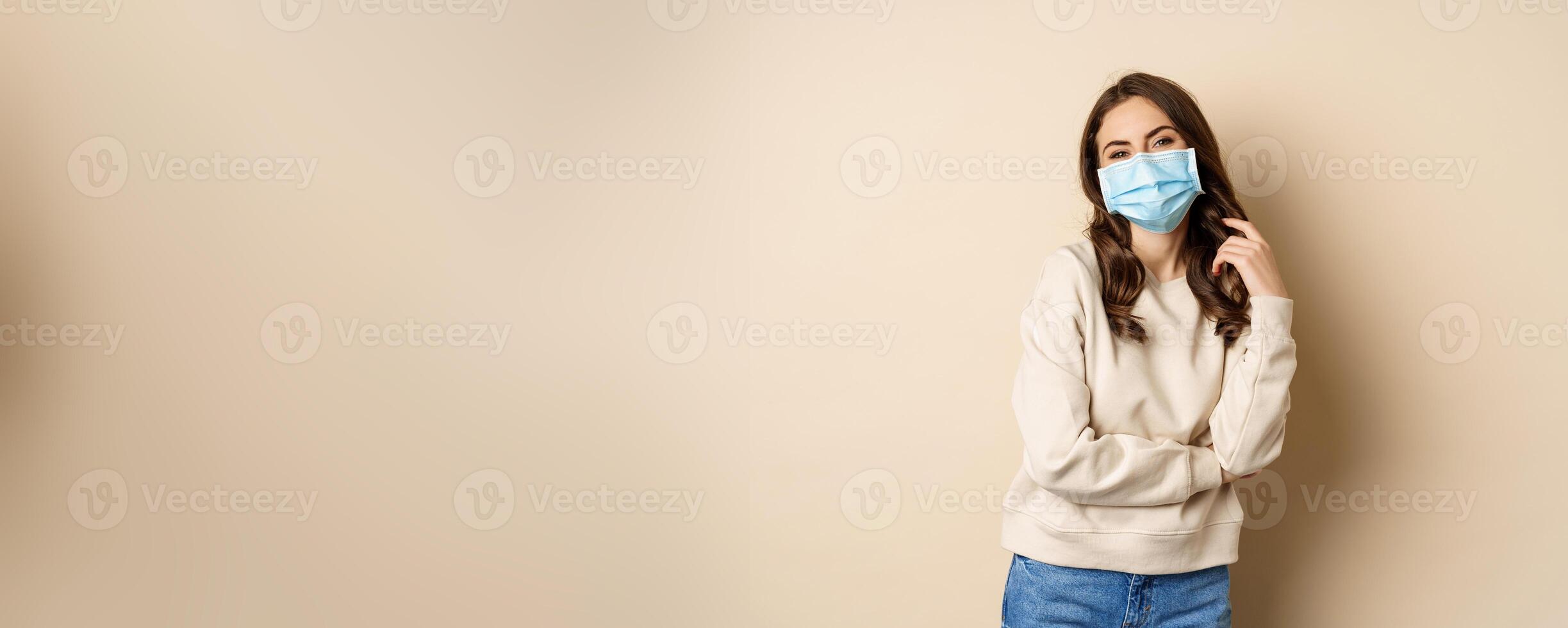 Covid-19, pandemic and quarantine concept. Beautiful modern woman in medical face mask, cross arms on chest, looking confident, beige background photo
