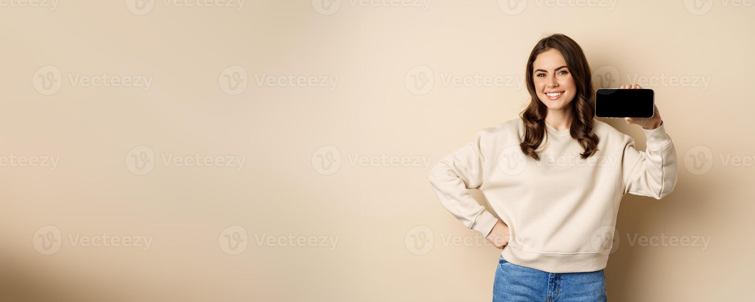 Portrait of smiling cute woman showing horizontal smartphone screen, website or application, standing over beige background photo