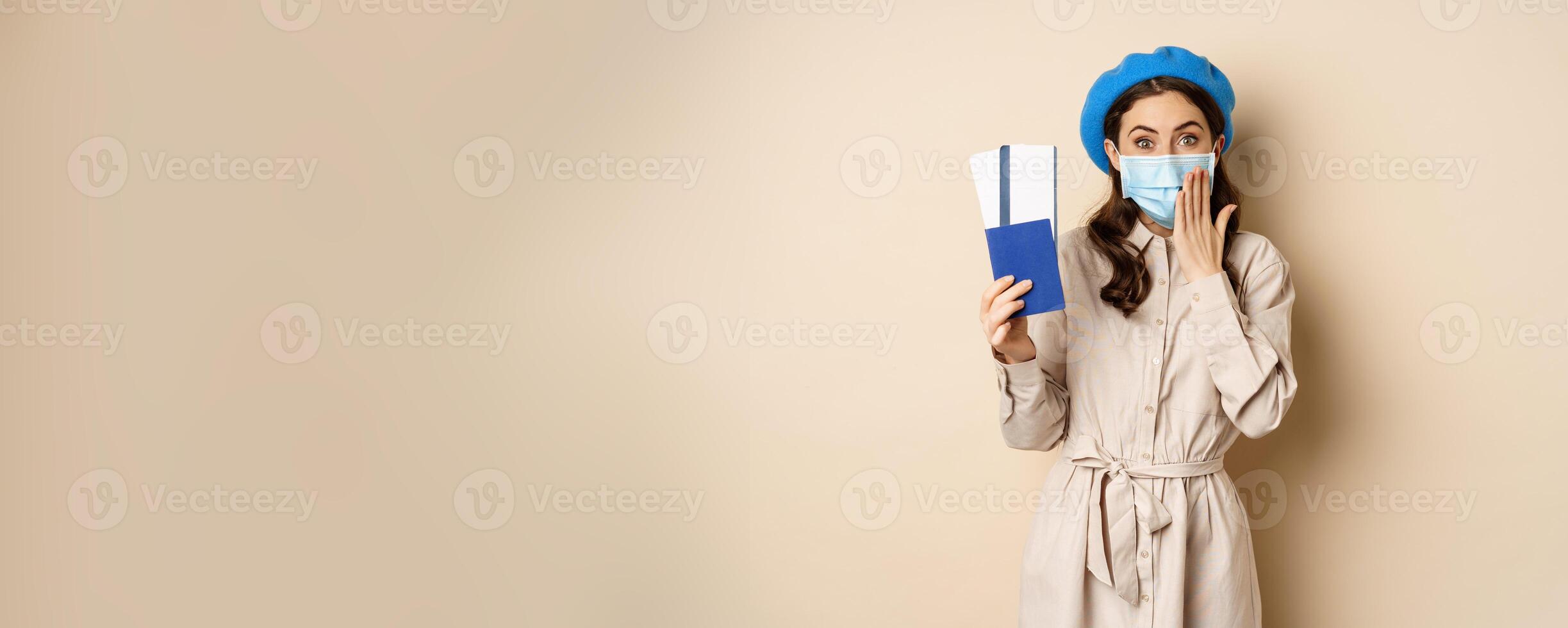 Covid pandemic and travelling concept. Portrait of cute girl in medical face mask going on trip, showing passport with tickets abroad and looking excited, beige background photo
