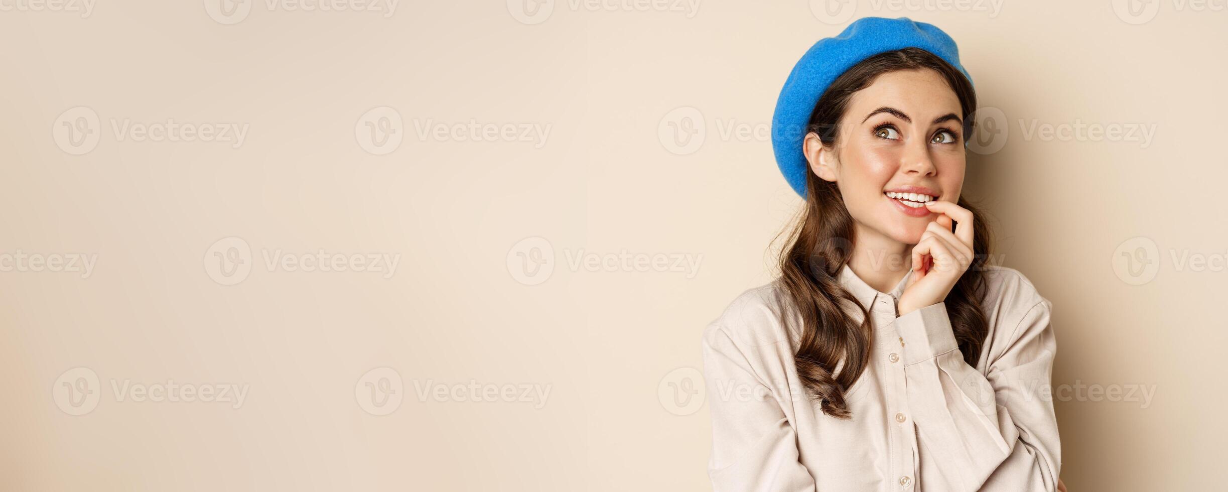 Close up portrait of dreamy smiling girl imaging, picturing smth, looking up and thinking, standing over beige background in blue trendy hat photo