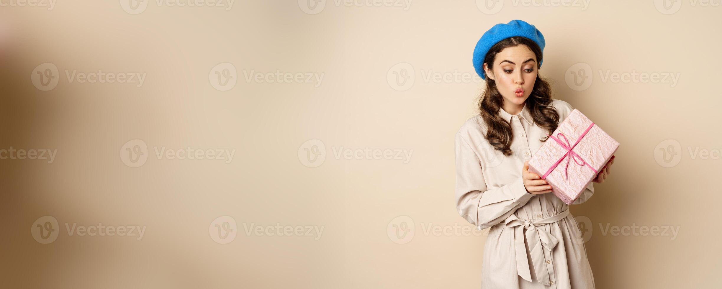 Holidays and gifts concept. Beautiful girl receive gift box and looking happy, holding pink wrapped present with joyful face expression, beige background photo