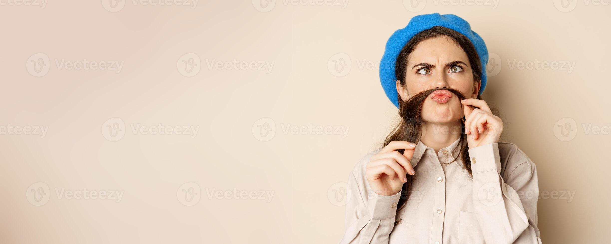 Funny beautiful girl fooling around, making moustache out of hair under lips, squinting and grimacing, standing happy against beige background photo
