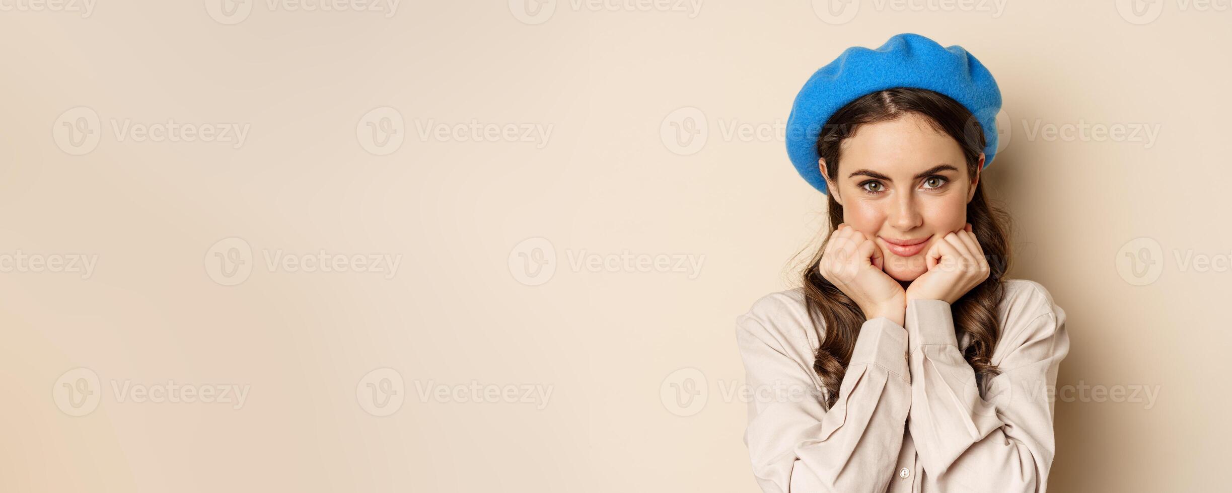 Close up portrait of feminine young woman in trendy french hat, posing cute and romantic, making silly coquettish face and tender smile, beige background photo