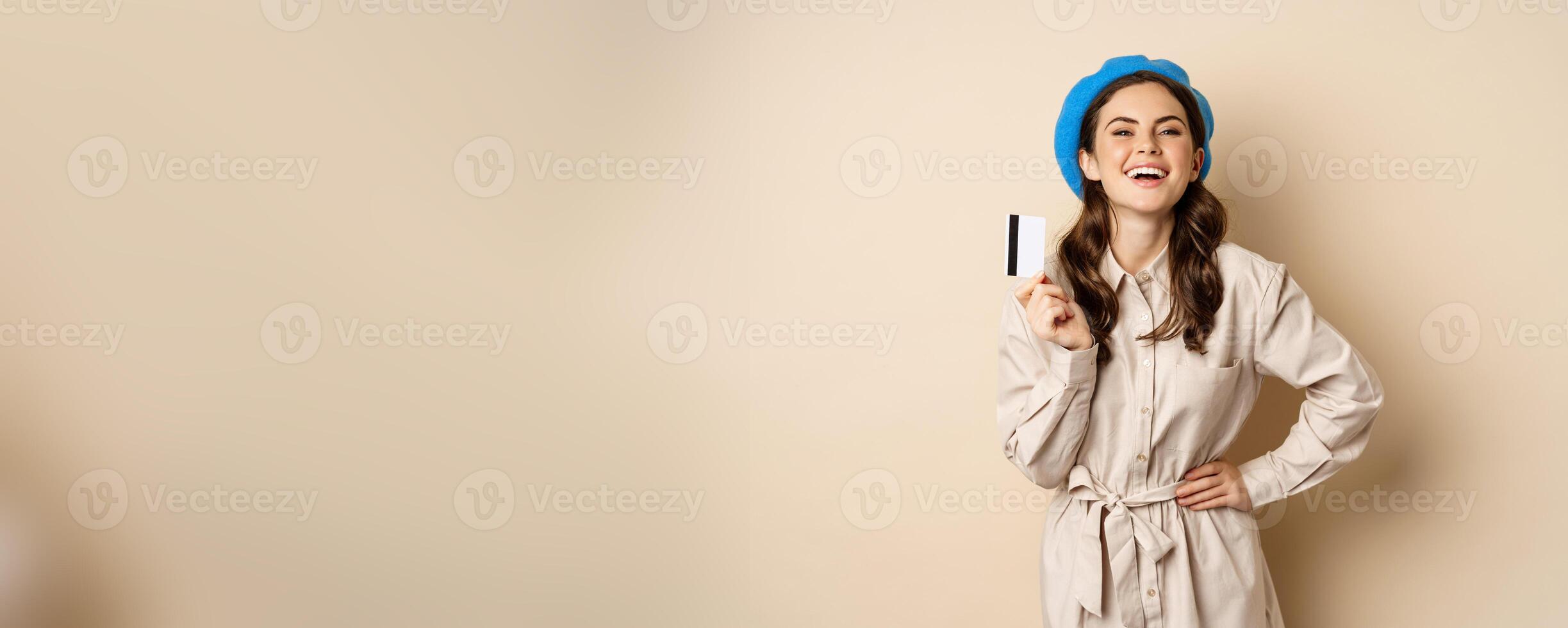Portrait of happy girl shopper, going on shopping and laughing, holding credit card, posing against beige background photo