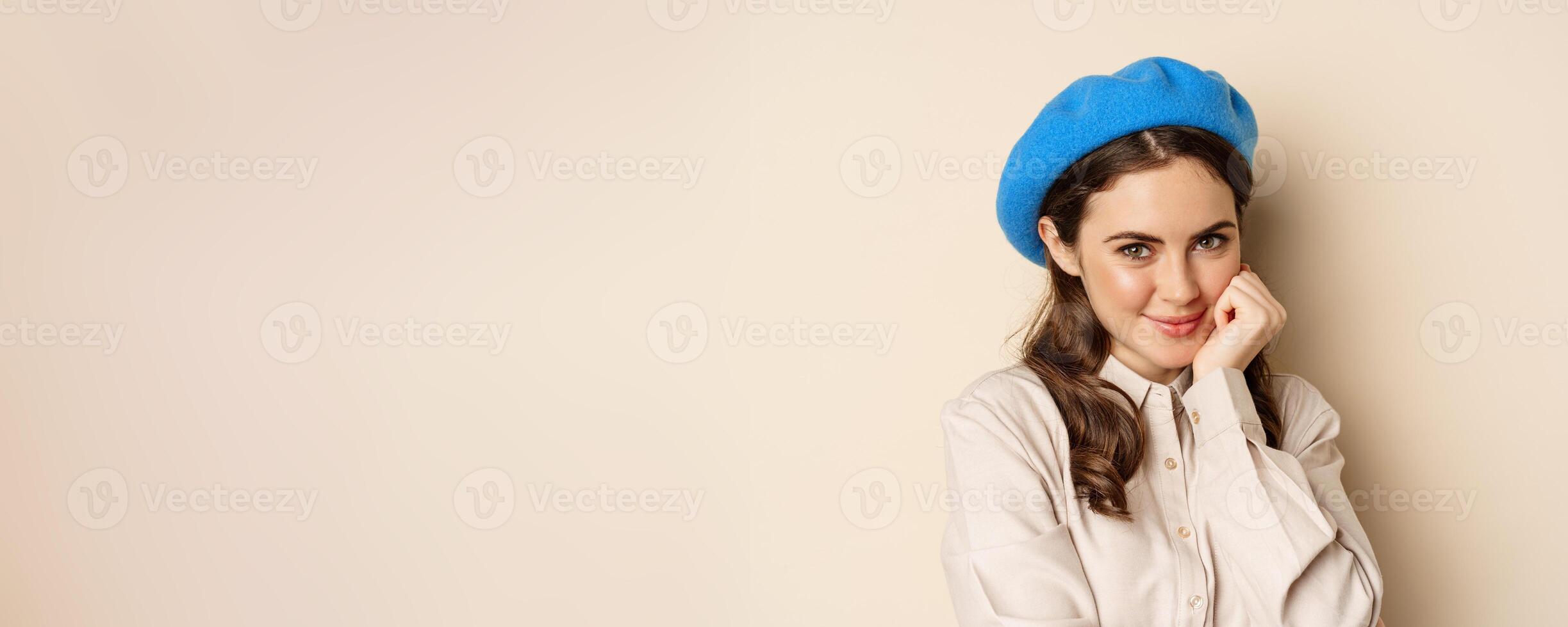 Close up portrait of feminine young woman in trendy french hat, posing cute and romantic, making silly coquettish face and tender smile, beige background photo