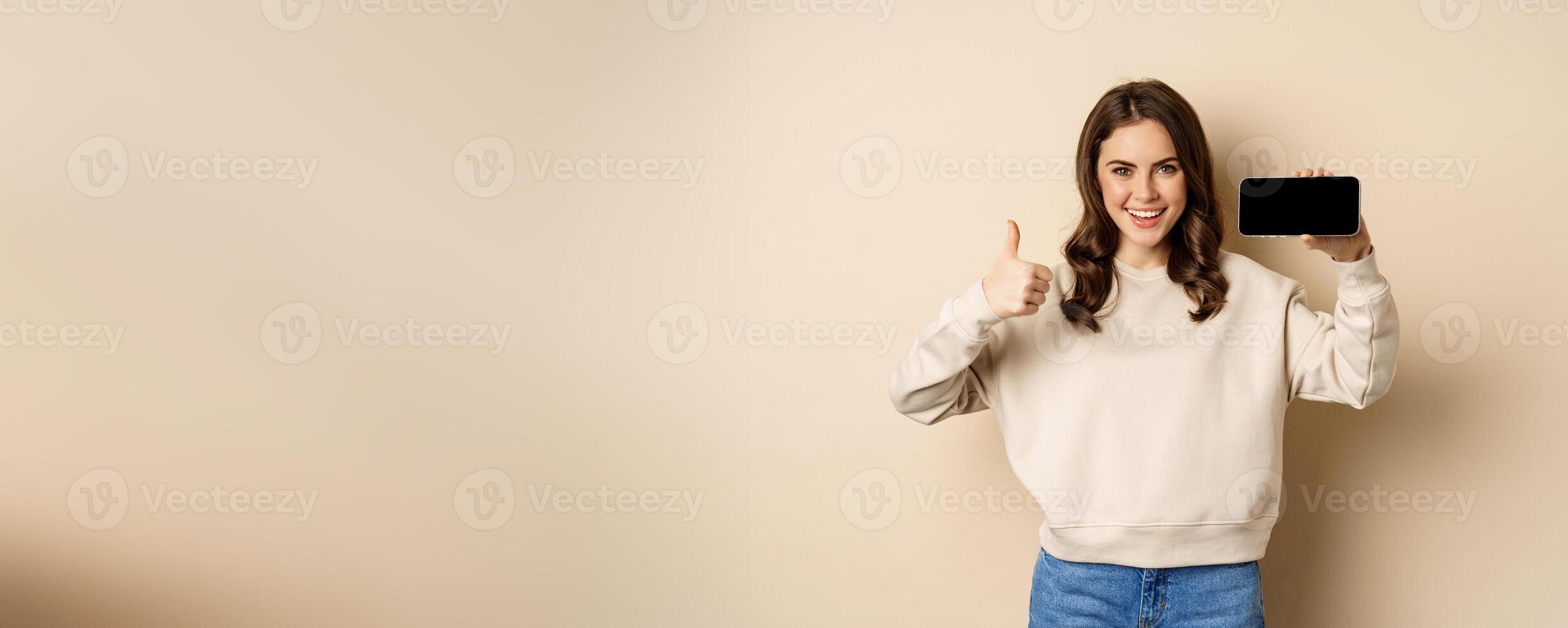 Smiling happy woman showing smartphone horizontal screen, thumbs up, recommending website, online store or app, standing over beige background photo