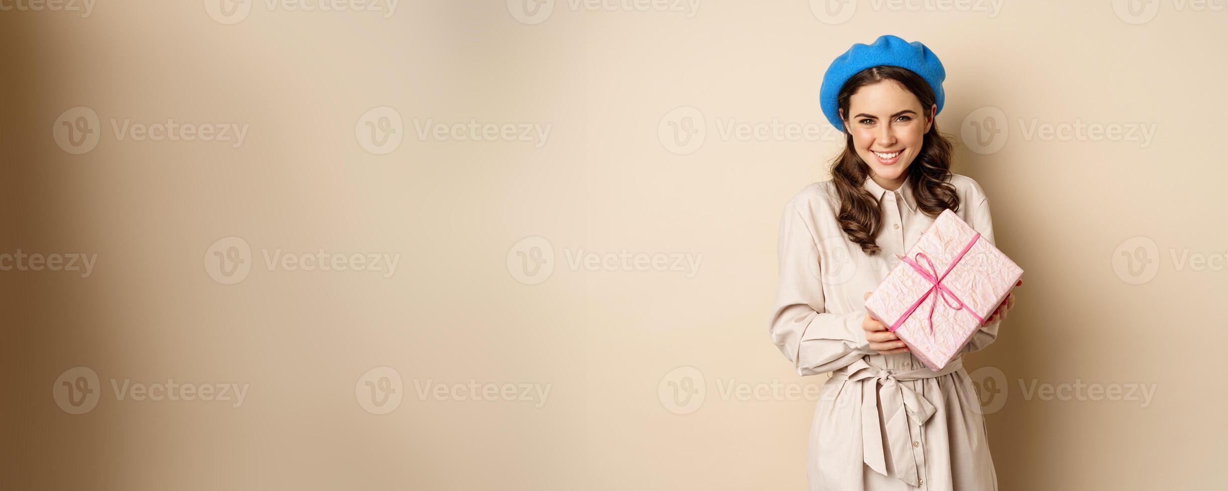 Holidays and gifts concept. Beautiful girl receive gift box and looking happy, holding pink wrapped present with joyful face expression, beige background photo