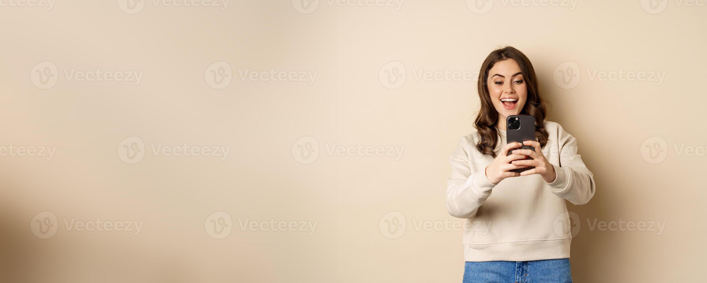 Happy woman recording , shooting photo on smartphone camera and smiling, standing over beige background