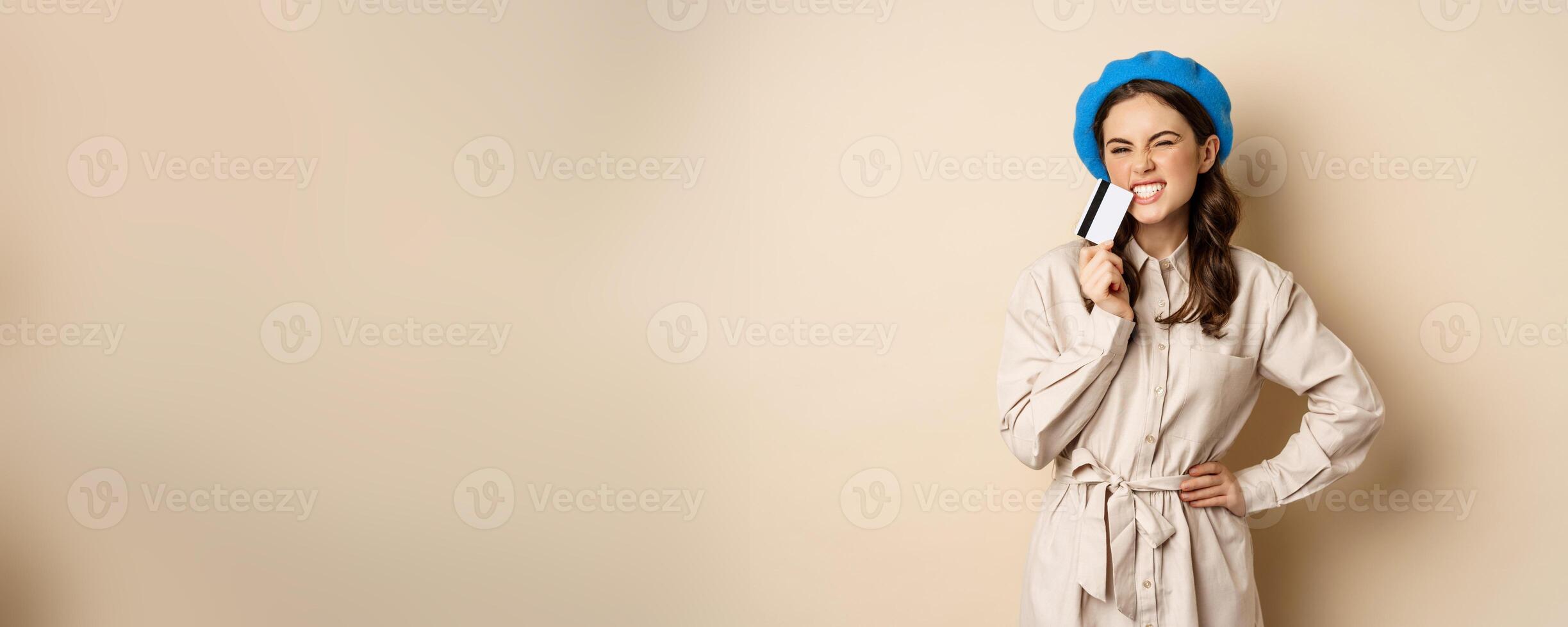 Shopping and discounts concept. Young stylish modern girl, posing with credit card, looking happy and smiling, standing over beige background photo