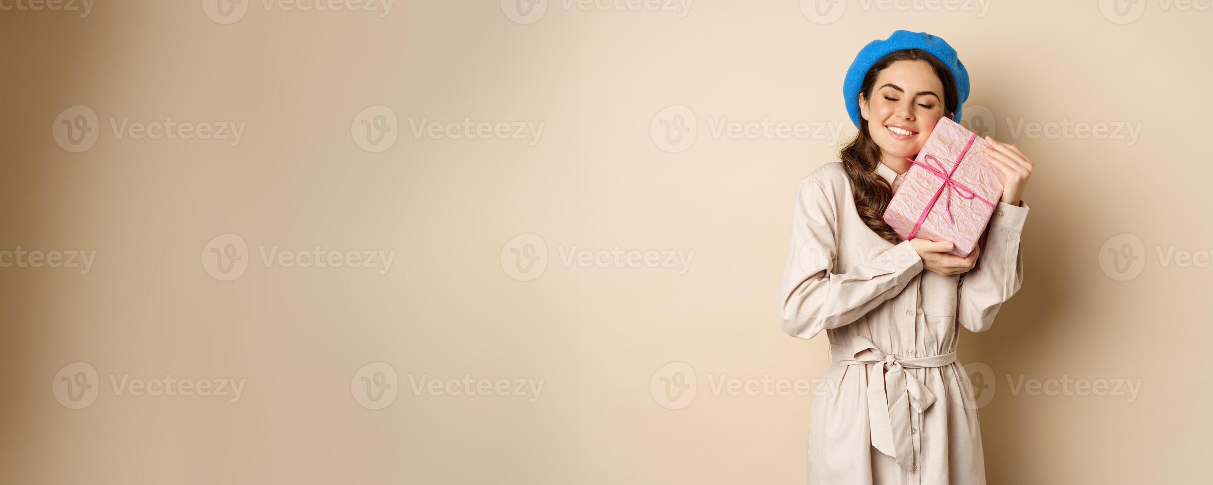 Holidays and gifts concept. Beautiful girl receive gift box and looking happy, holding pink wrapped present with joyful face expression, beige background photo