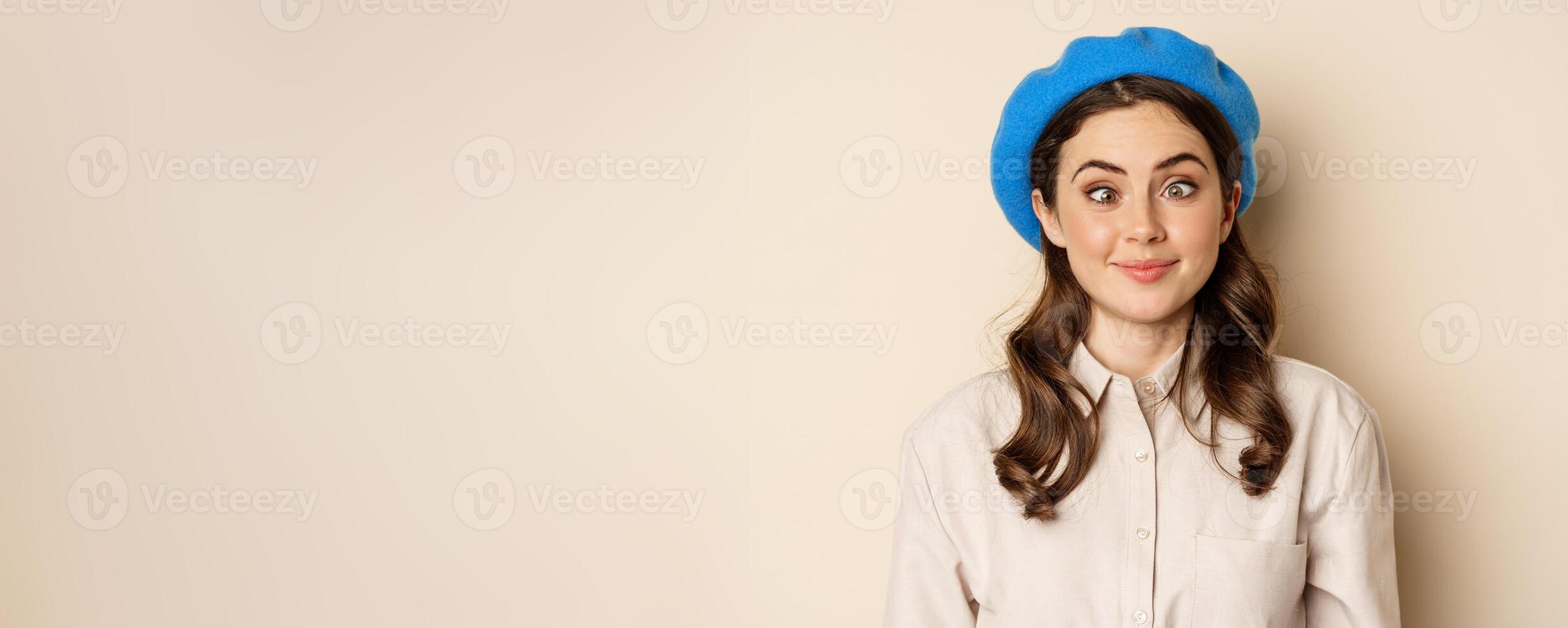 Close up portrait, silly face of young funny girl, squinting eyes and having fun, posing in trench and hat against beige background photo