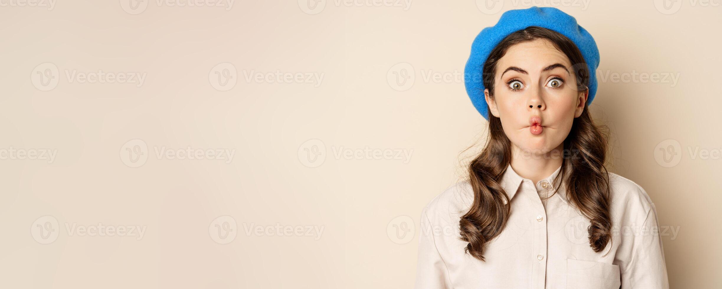 Young funny woman acting, fooling around, pucker lips and grimacing, posing against beige background in trendy blue hat photo