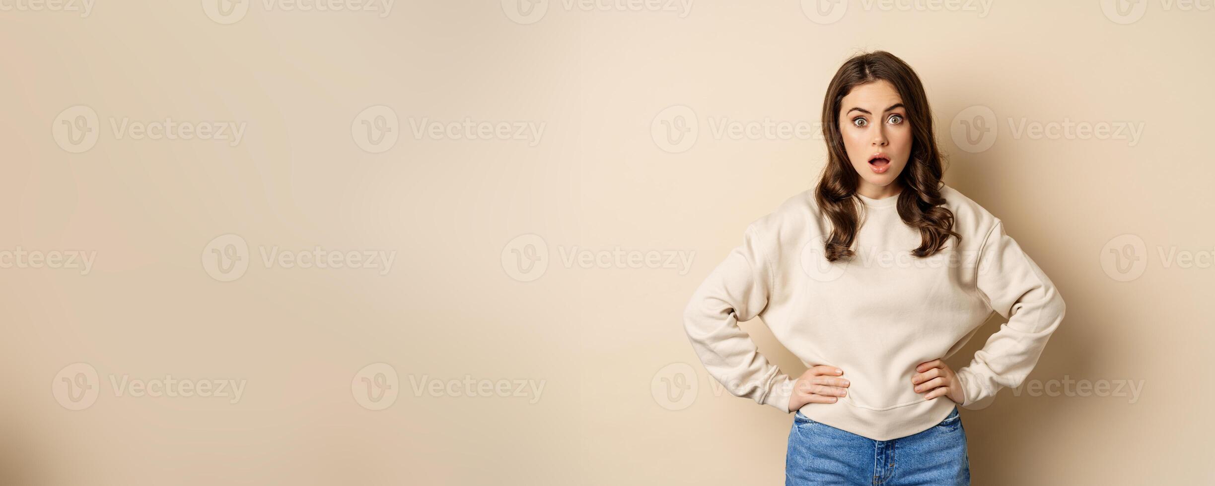Portrait of shocked brunette woman drop jaw, gasping and staring speechless at camera, beige background photo