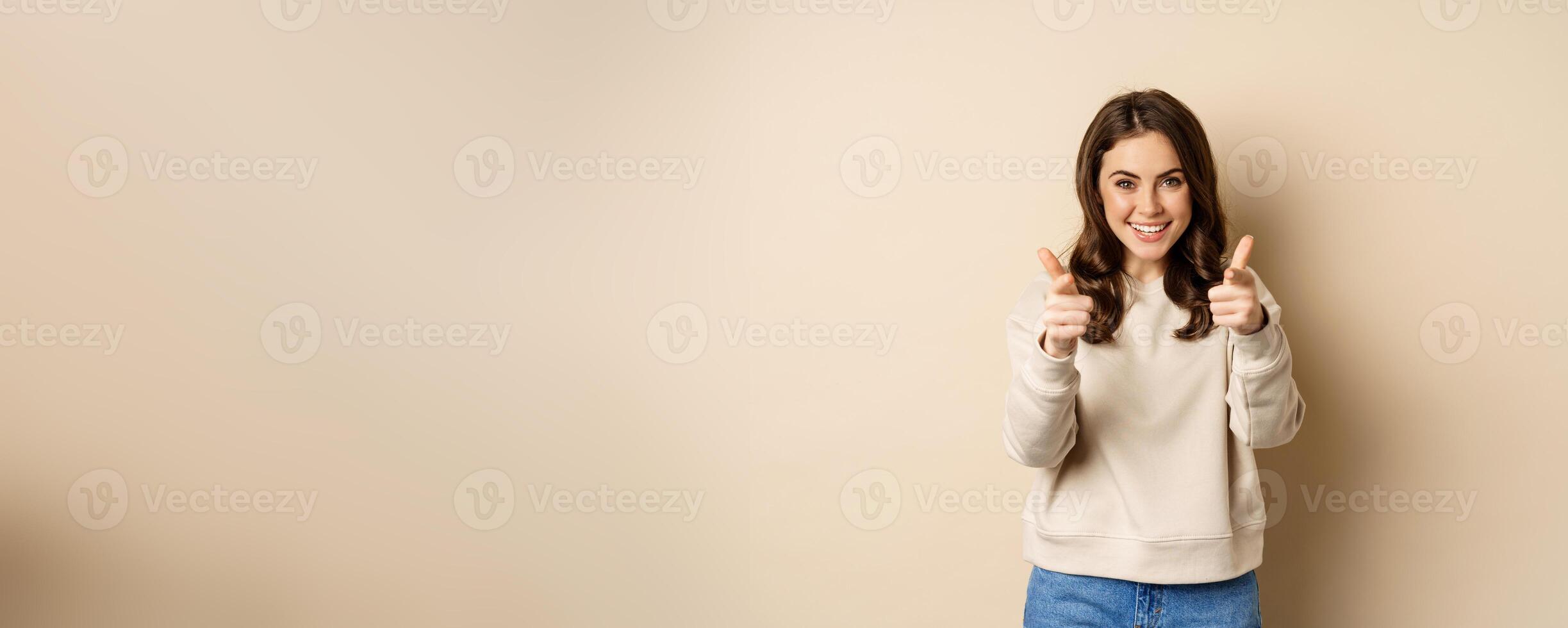 Smiling happy woman choosing you, pointing fingers at camera and laughing, inviting people, standing over beige background photo