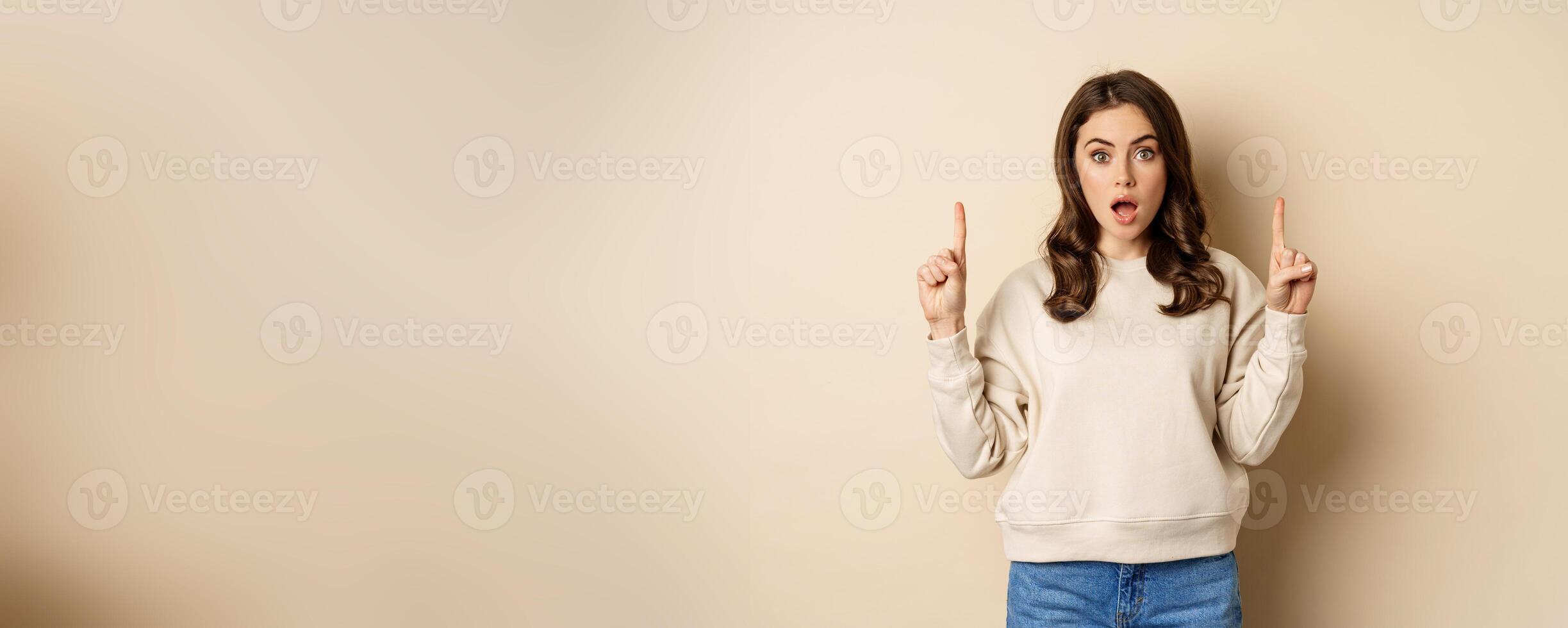 Surprised beautiful girl showing advertisement, pointing fingers on top, store sale or website link up, standing over beige background photo