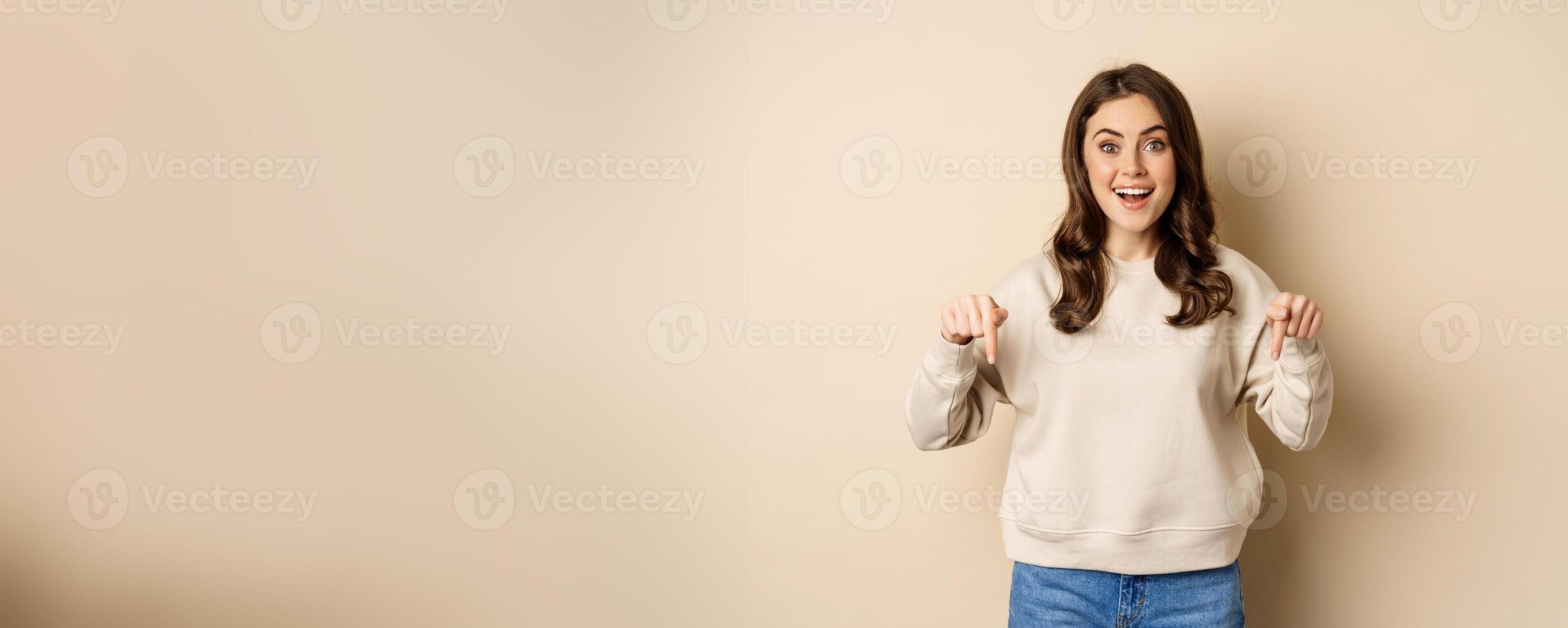 Enthusiastic attractive girl pointing fingers down, showing advertisement, link or promo below, standing over beige background photo