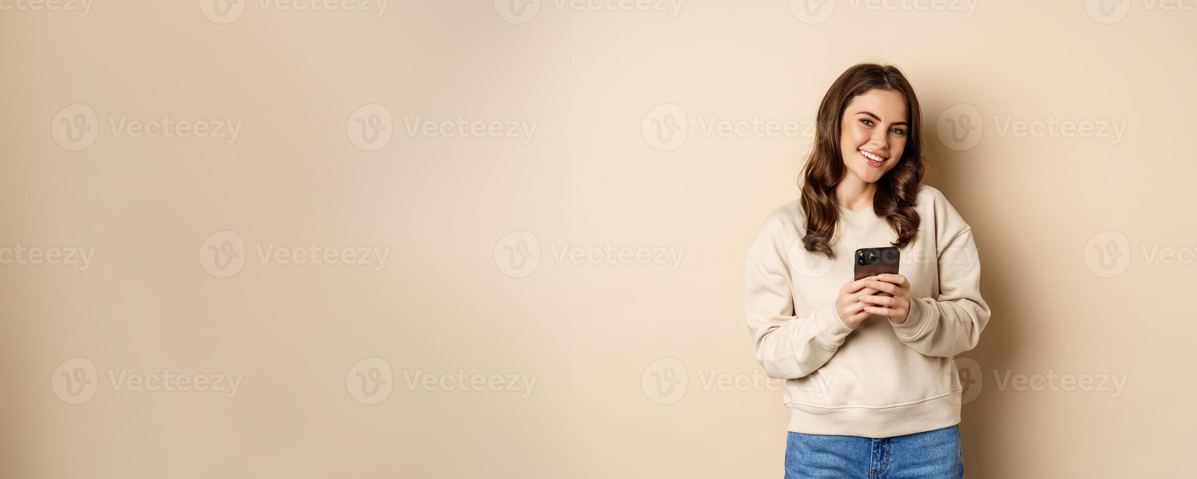 People and cellular technology. Beautiful stylish woman using mobile phone, smartphone app, smiling and looking at screen, standing over beige background photo