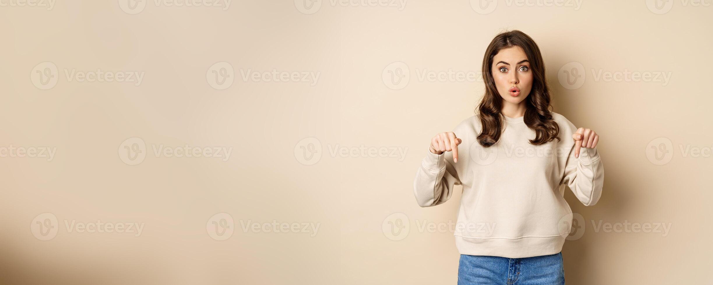 Enthusiastic attractive girl pointing fingers down, showing advertisement, link or promo below, standing over beige background photo