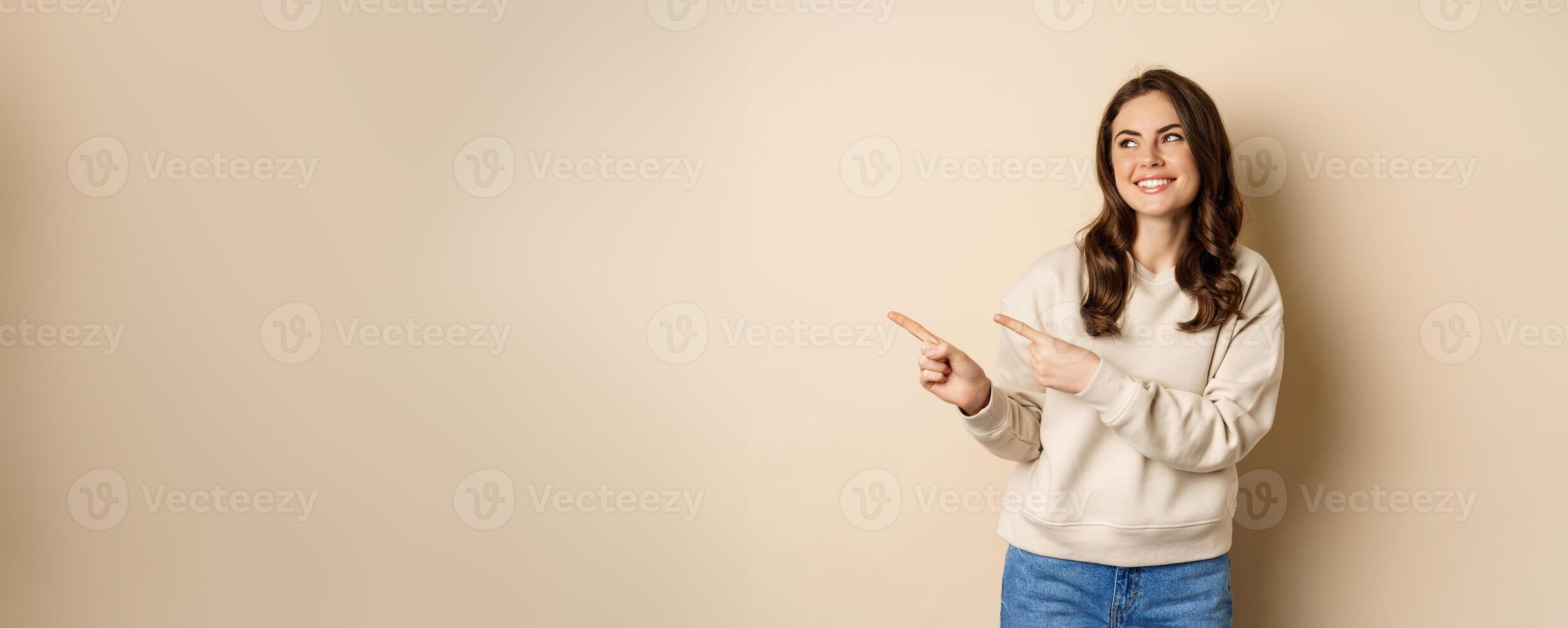 Beautiful girl pointing and looking left, laughing and smiling happy while showing advertisement, logo or store banner, beige background photo