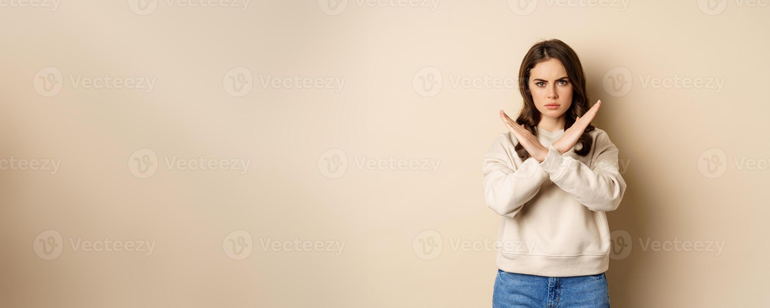 Woman showing stop, cross sign gesture and frowning angry, dislike and disapprove, saying no, rejecting and prohibiting, standing over beige background photo