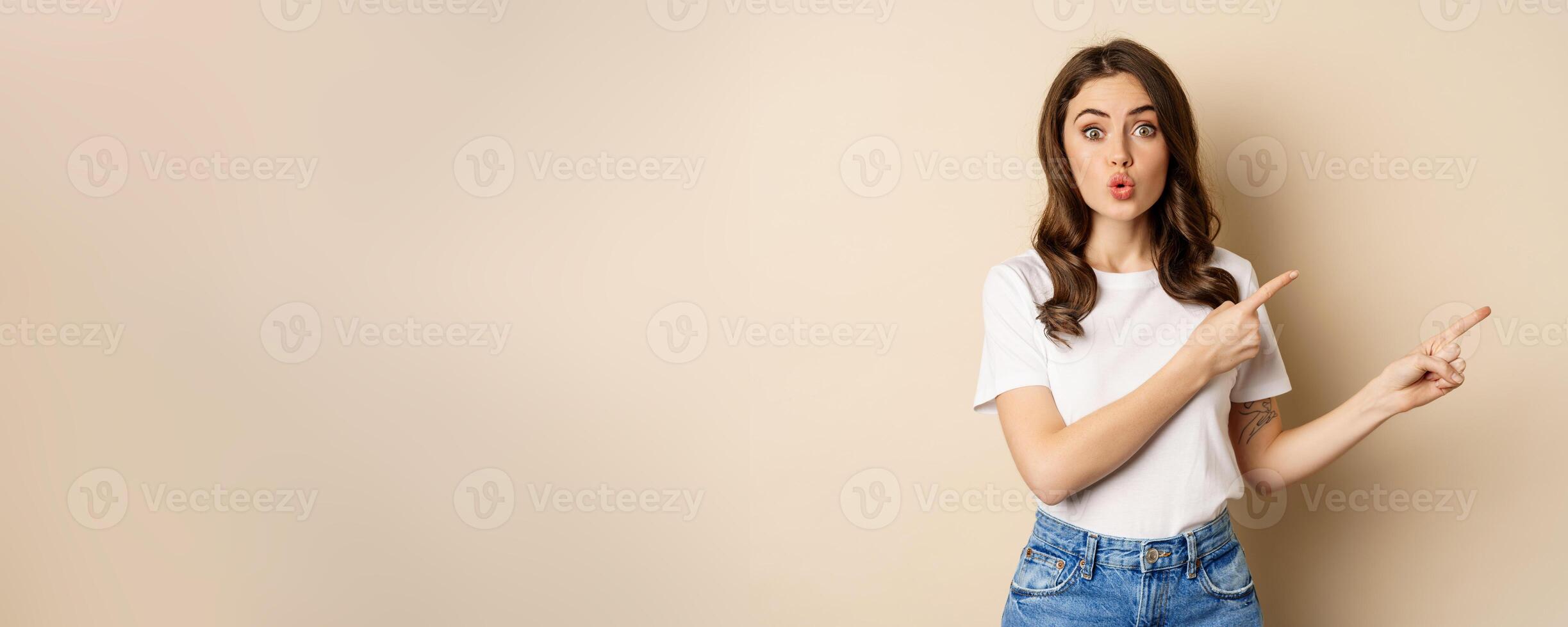 Excited girl pointing fingers right, showing promo with curious face expression, standing over beige background photo