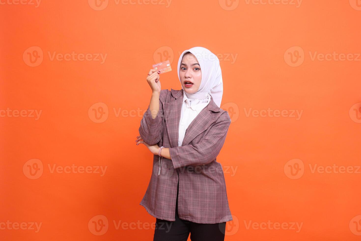 Young indonesia woman in hijab suit surprised with arms crossed while showing isolated plastic debit credit card on orange background. customer satisfied with service for financial, mobile transaction photo