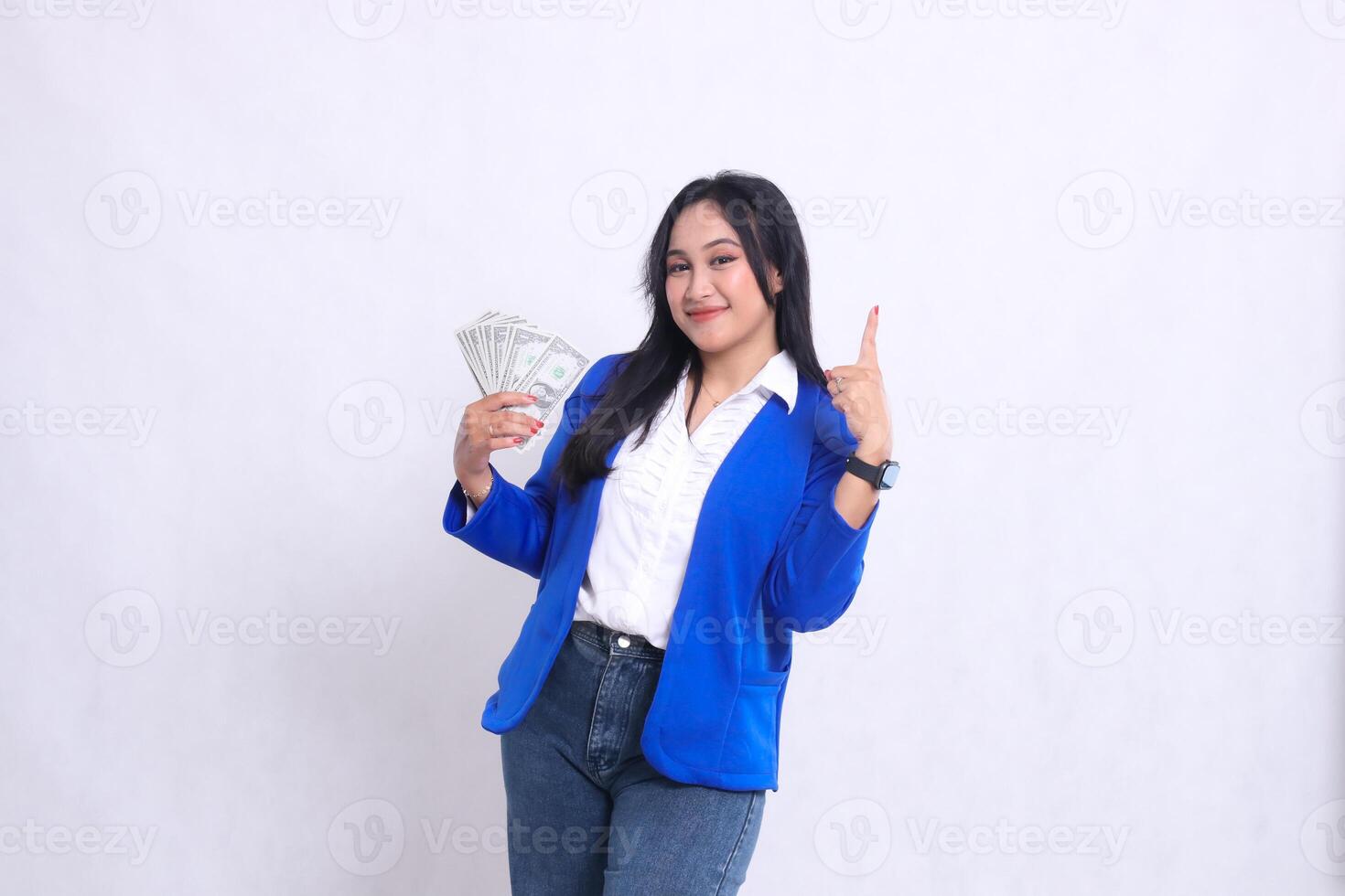 beautiful adult office girl wearing blue formal suit standing smiling to camera holding dollar bill to right and pointing up isolated white background. for transaction, finance, business content photo