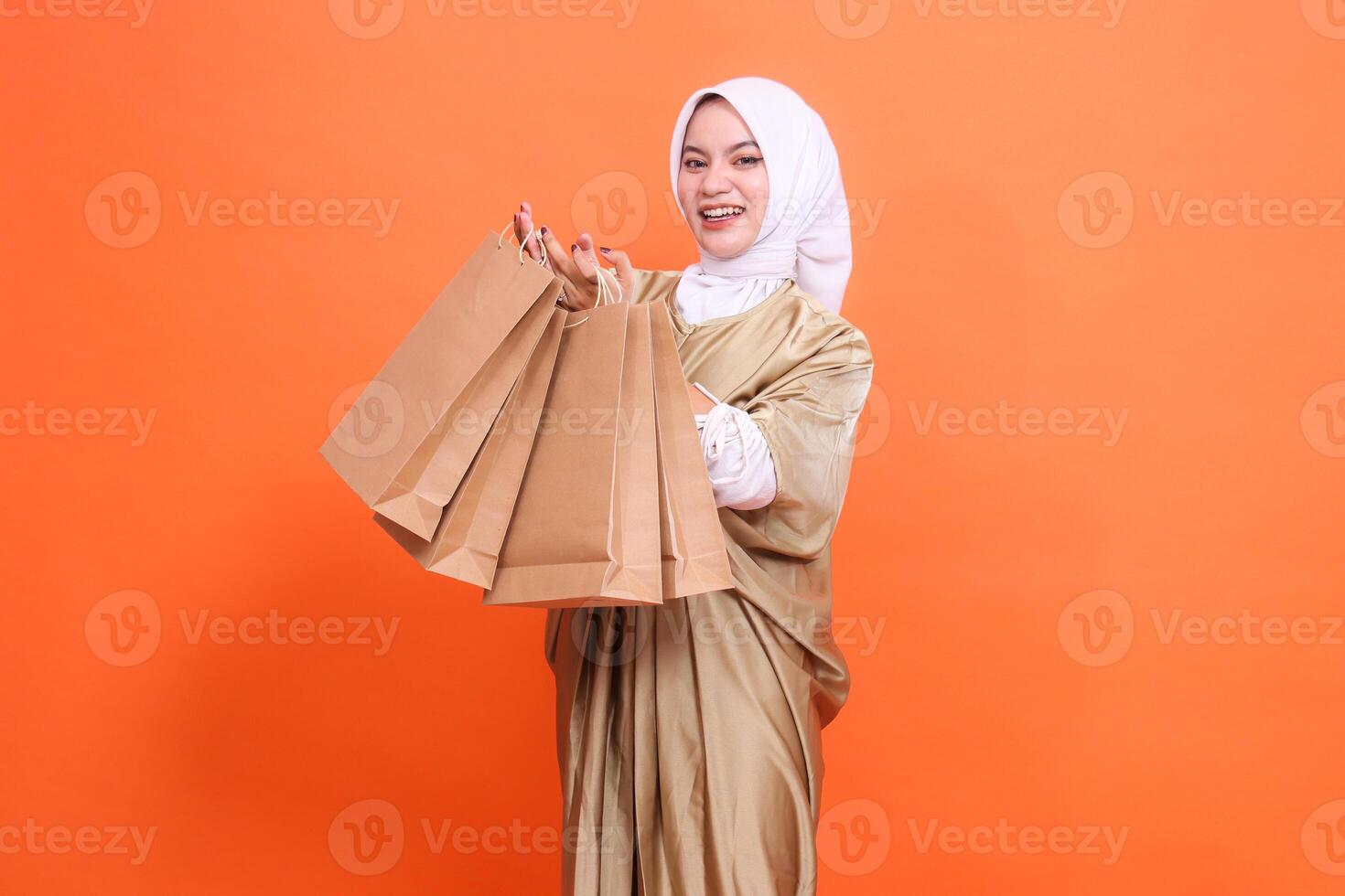 Happy young Asian woman with both hands carrying a brown paper bag containing groceries in front of her wearing a kaftan Muslim dress with a hijab, isolated on an orange background. Concept of fashion photo