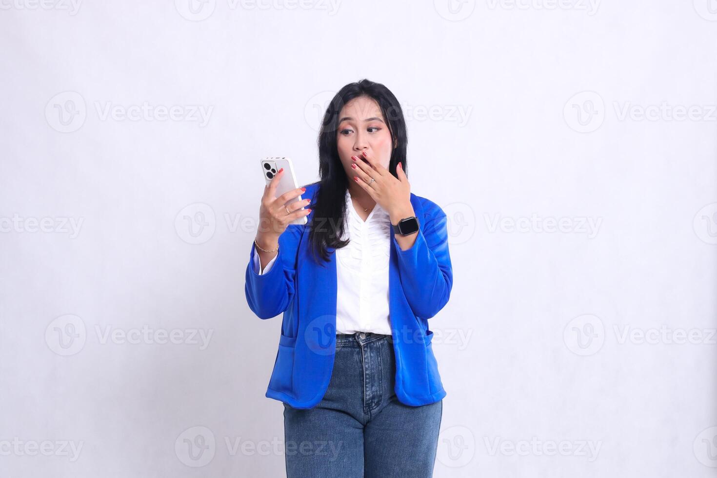 beautiful young office girl wearing blue formal suit standing shocked candid operating smartphone handphone while her hand covers her mouth. for technology content, lifestyle, mobile transaction photo