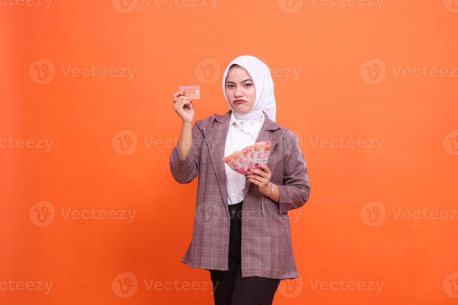 Young indonesia woman in her 20s frowning wearing hijab office wear holding debit credit card and money isolated on plain pastel orange background studio portrait photo