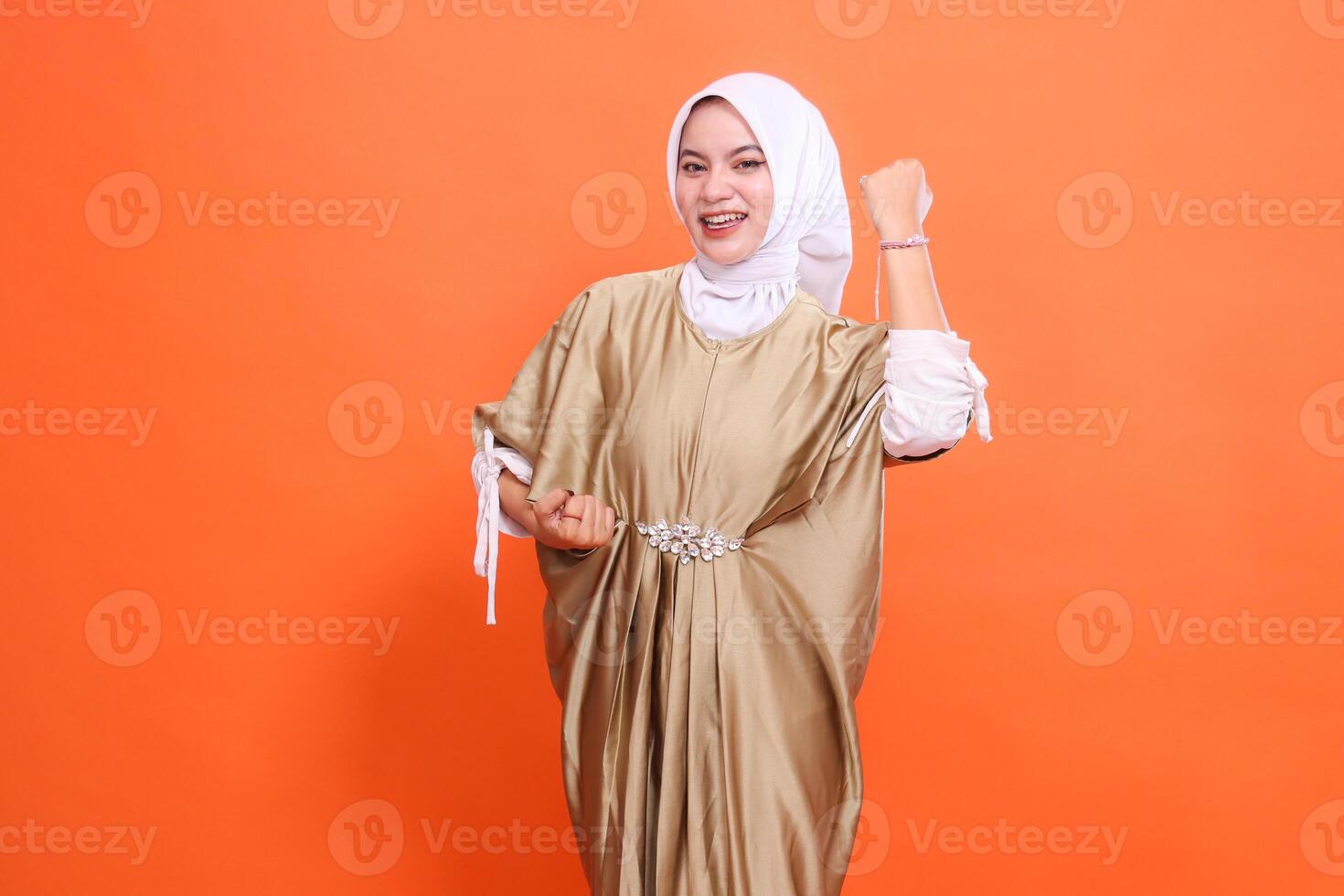 beautiful adult indonesia woman happy with both hands clenched up and down successfully wearing a kaftan Muslim dress with a hijab, isolated on an orange background. stylish fashion, promotion photo