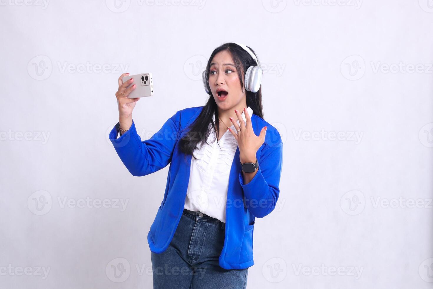 beautiful mature office woman wearing blue formal suit standing on right side shocked losing playing on smartphone mobile phone and headphone isolation white background. for entertainment content photo