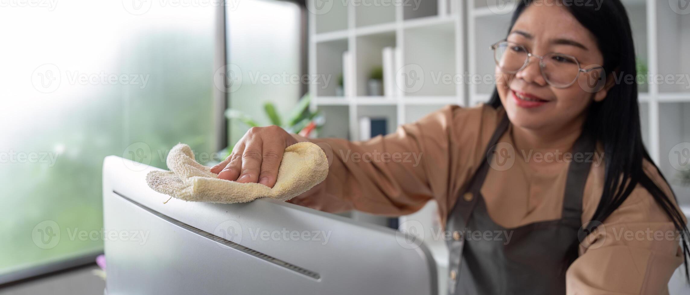 Professional Housekeeper Cleaning a Modern Home Office with a Smile, Wearing Apron and Glasses, Using a Cloth to Wipe Down Surfaces photo