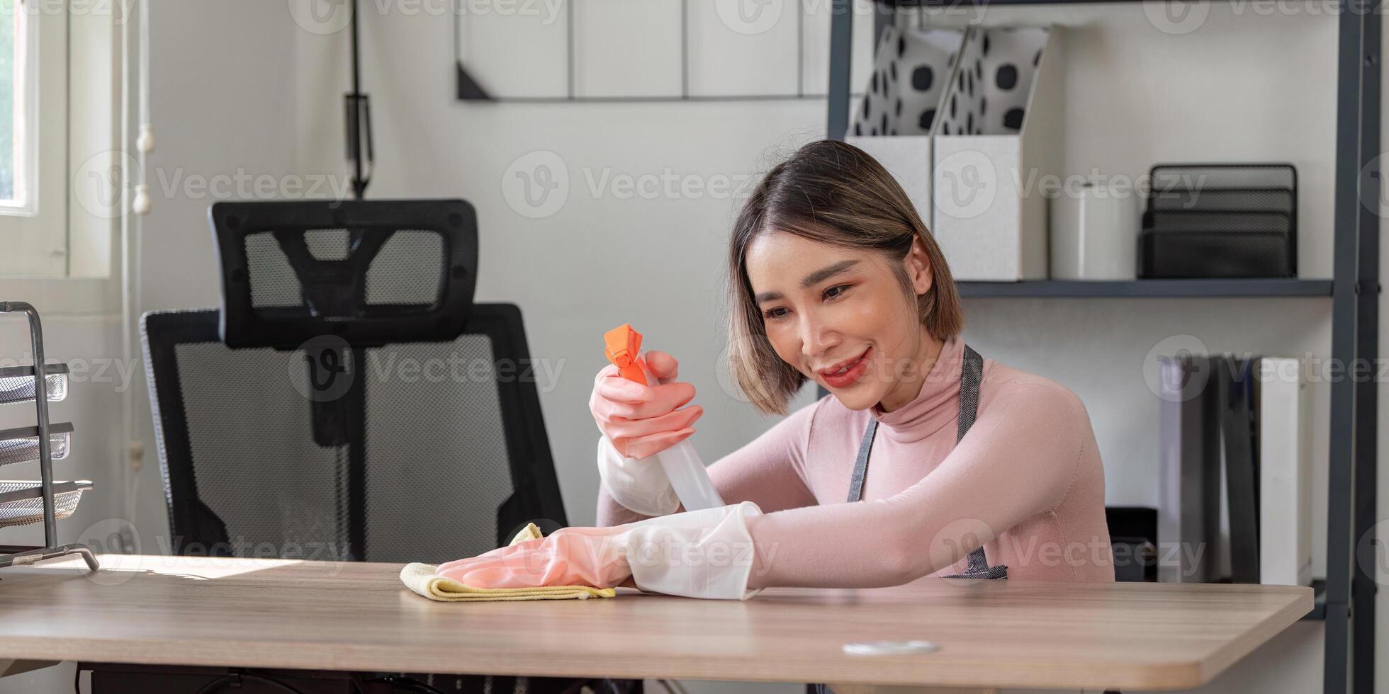Young Woman Cleaning Desk in Modern Home Office with Spray Bottle and Cloth, Smiling and Focused on Task photo