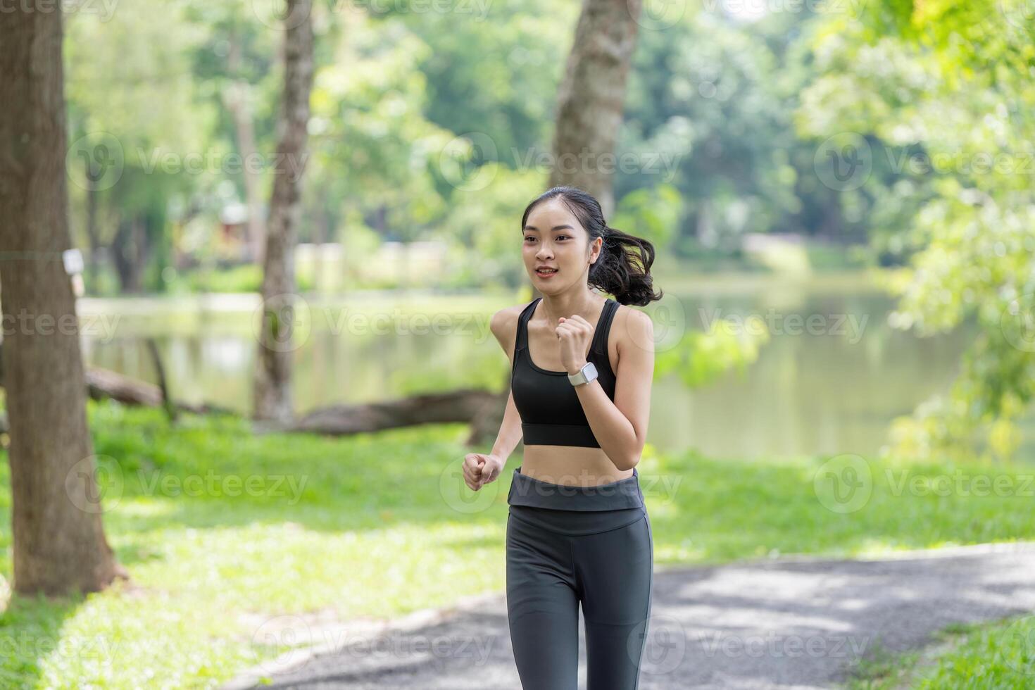 Young Woman Jogging in a Park on a Sunny Day, Wearing Sportswear and a Smartwatch, Surrounded by Lush Greenery and Trees photo