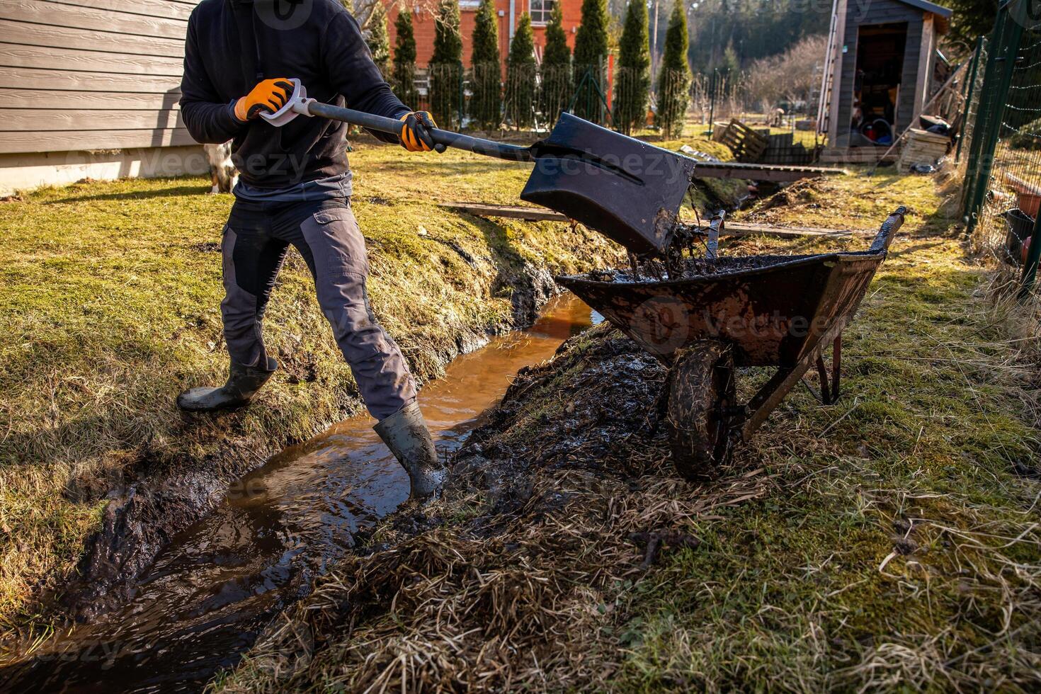 Man in protective gear tirelessly working to dredge a muddy trench with a shovel and wheelbarrow in a backyard during early spring photo