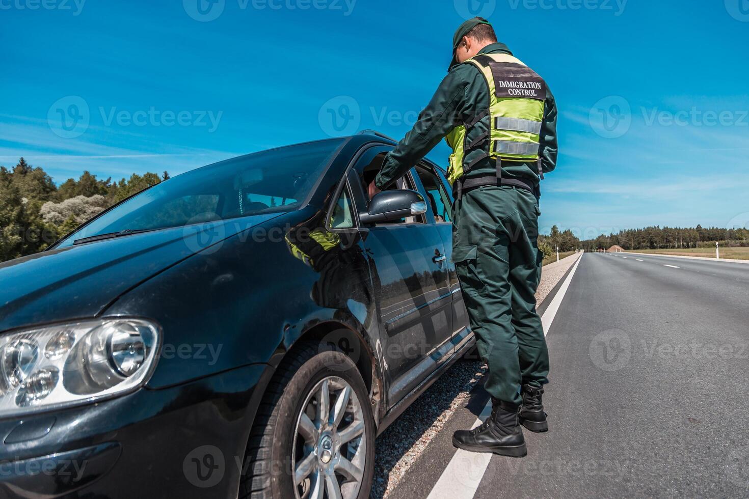 border patrol officer inspecting vehicle at checkpoint on sunny day with clear skies and long open road photo