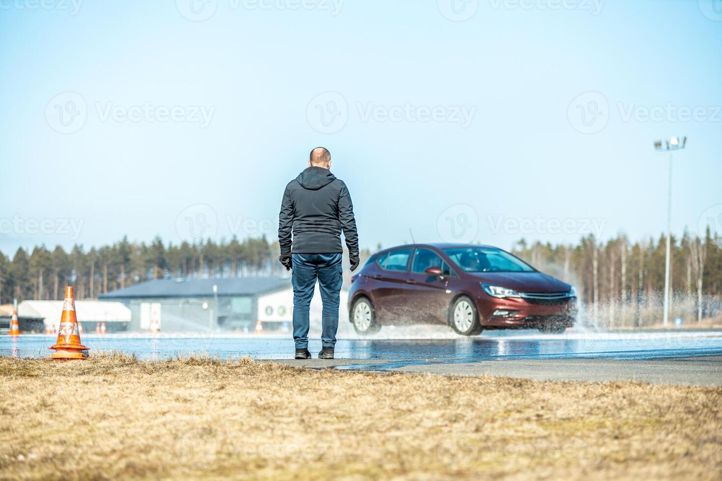 man observing a wet road test with a maroon car skidding on a water-covered track, testing vehicle safety and tire performance under controlled conditions photo
