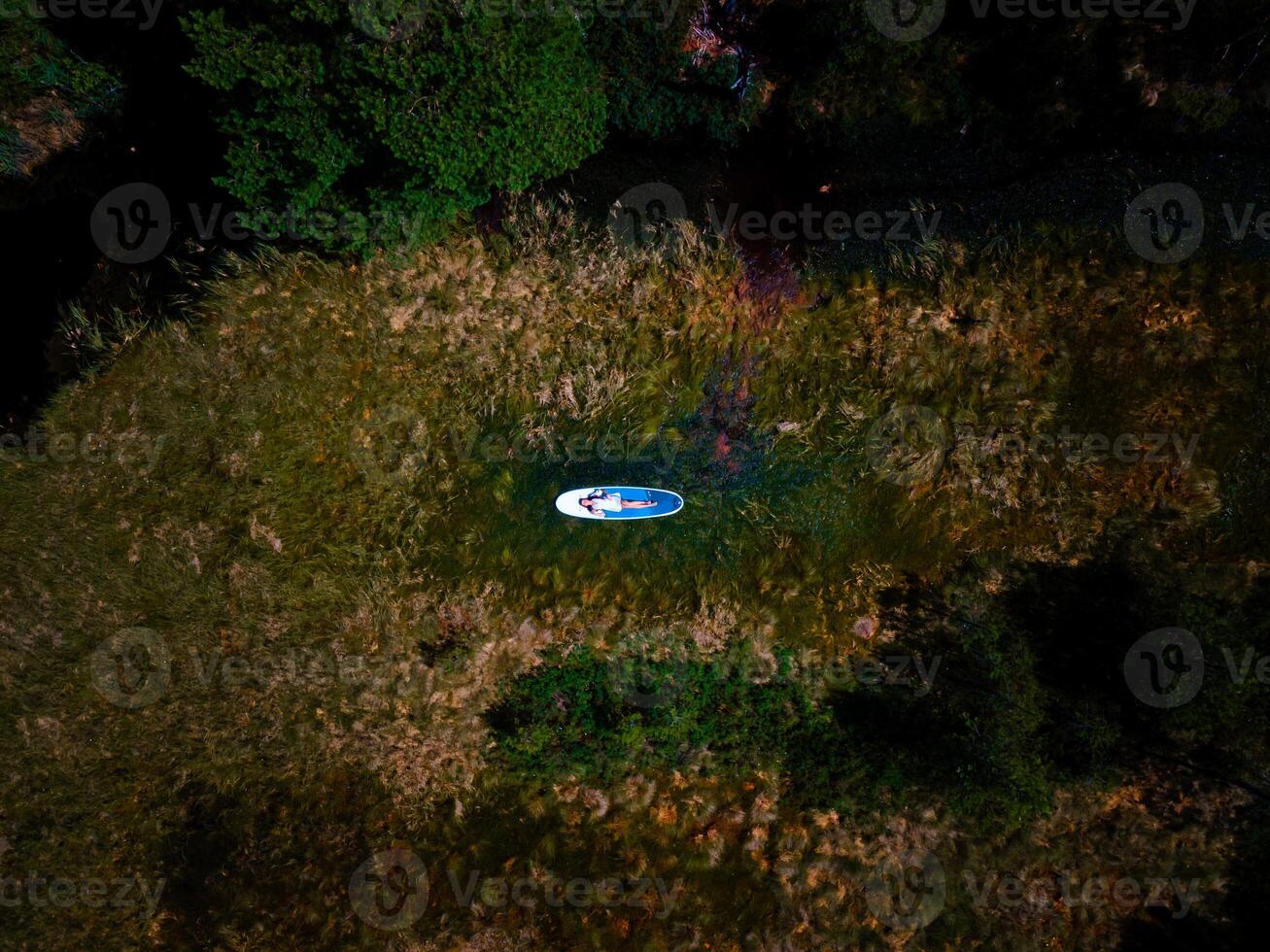 A beautiful woman in a white dress is lying on a paddle surfboard and relaxing in the swamp photo